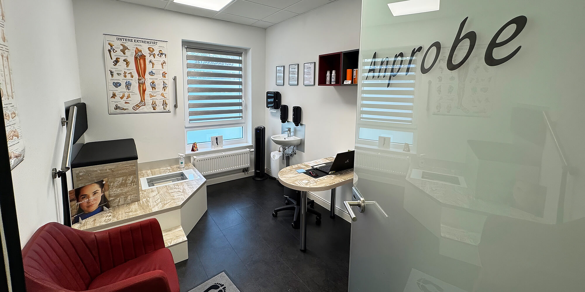 Anproberaum mit Schreibtisch, Waschbecken, anatomischem Poster und Glaswand mit der Aufschrift Anprobe